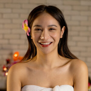 Smiling Asian woman with a flower in her hair, wrapped in a towel, promoting confidence and relaxation in a body sculpting setting.
