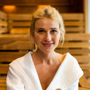 Woman relaxing in a spa, wearing a white robe, smiling, surrounded by wooden decor, representing confidence and relaxation in body sculpting treatments.